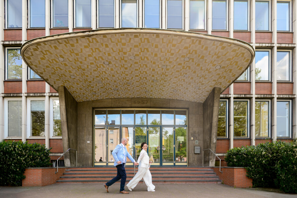 braut und Bräutigam vor dm Standesamt. Braut gehr voran und hält Bräutigam an der Hand